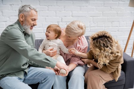 Téléchargez les photos : Tout-petit fille souriant près des grands-parents et de la mère s'amuser sur le canapé dans le salon - en image libre de droit