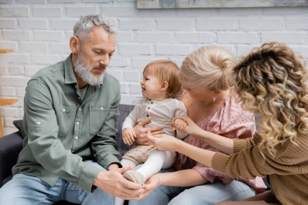 mature couple touching leg of cheerful granddaughter while sitting on sofa in living room