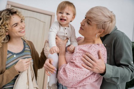 fröhliche Frau schaut Kleinkind Tochter lächelnd in die Hände der Großmutter in Flur der Wohnung