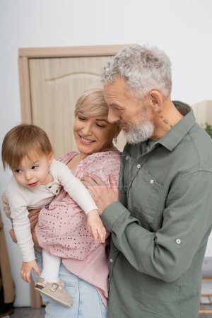 Jubelnde Frau umarmt kleine Enkelin neben bärtigem Ehemann im Hausflur
