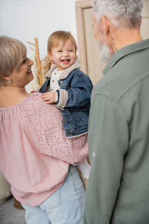 middle aged woman holding cheerful granddaughter in denim jacket near family at home