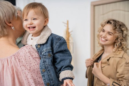 happy child in denim jacket smiling in hands of granny near blurred mother at door of apartment