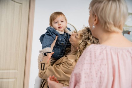 Téléchargez les photos : Femme d'âge moyen floue regardant sa fille tenant une petite fille dans une veste en denim près de la porte d'entrée de l'appartement - en image libre de droit