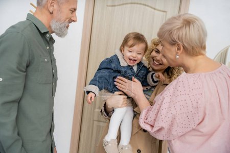 overjoyed child in denim jacket smiling near mother and grandparents near entrance door at home