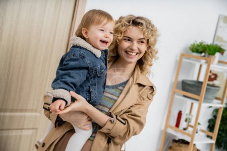 joyful blonde woman with wavy hair holding carefree daughter in denim jacket near entrance door at home