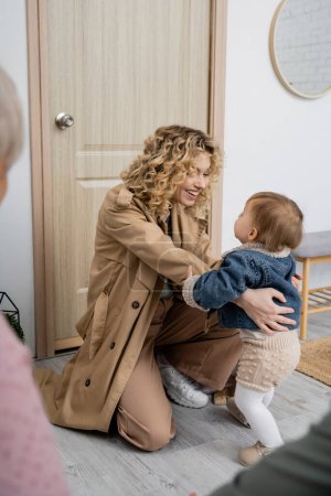 cheerful woman in trench coat embracing little daughter near entrance door at home
