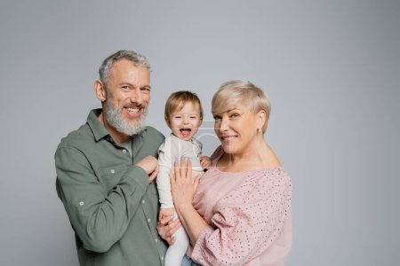 glückliches älteres Paar blickt in die Kamera neben lachender Enkelin isoliert auf grau