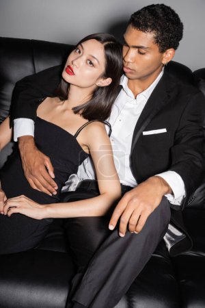 Téléchargez les photos : Sensuelle asiatique femme avec rouge lèvres regarder caméra près élégant afro-américain gars sur canapé noir sur fond gris - en image libre de droit
