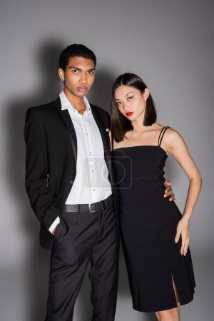 Téléchargez les photos : Afro-américain homme en costume noir et asiatique femme en robe de sangle regardant caméra sur fond gris - en image libre de droit