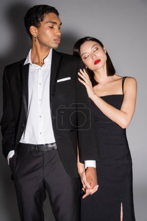 elegante mujer asiática con los ojos cerrados apoyándose en elegante hombre afroamericano en traje negro sobre fondo gris