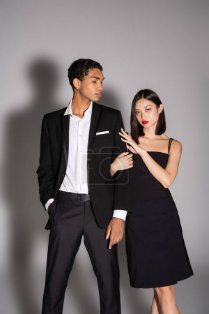 Téléchargez les photos : Brunette asiatique femme en robe noire regardant caméra près élégant afro-américain homme debout avec la main dans la poche sur fond gris - en image libre de droit