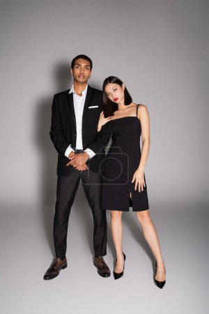 Photo for Full length of interracial couple in elegant formal wear looking at camera on grey background - Royalty Free Image