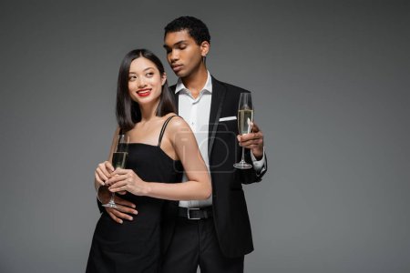 Téléchargez les photos : Souriant asiatique femme et élégant afro-américain tenue champagne verres isolés sur gris - en image libre de droit