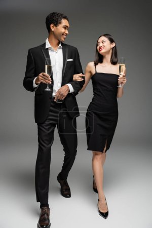 Téléchargez les photos : Pleine longueur de couple interracial heureux et élégant marchant avec des verres de champagne et se regardant sur fond gris - en image libre de droit