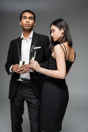 Téléchargez les photos : Jeune couple interracial en noir élégant vêtements posant avec des verres de champagne isolé sur gris - en image libre de droit