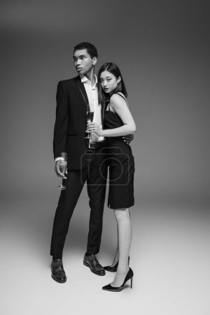 full length of interracial couple in elegant and stylish attire standing with champagne glasses on grey background