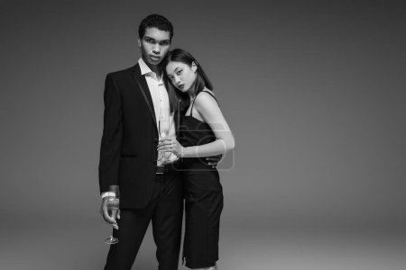 joven pareja interracial en elegante ropa formal posando con copas de champán aisladas en gris