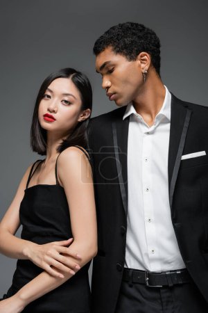 asiatische Frau in schwarzem Trägerkleid posiert in der Nähe junger afrikanisch-amerikanischer Mann in elegantem Blazer isoliert auf grau