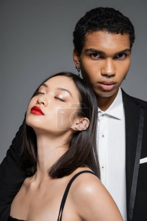 elegante africano americano hombre mirando cámara cerca sensual asiático mujer con rojo labios y cerrado ojos aislado en gris