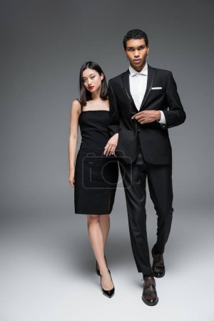 longitud completa de la pareja multiétnica en elegante ropa formal caminando y mirando a la cámara sobre fondo gris