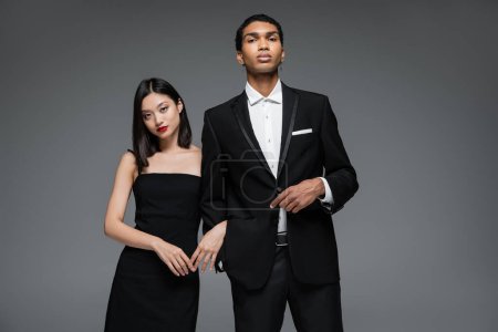 young interracial couple in elegant formal wear looking at camera isolated on grey