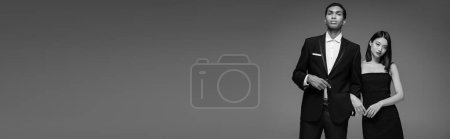 Photo for Young interracial models in black elegant clothes looking at camera isolated on grey, banner - Royalty Free Image