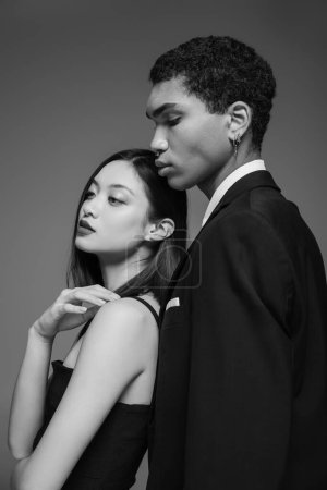sensual asian woman looking away near elegant african american man in black blazer isolated on grey