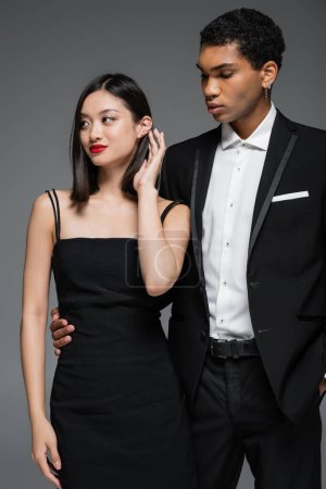 Afrikanisch-amerikanischer Mann im schwarzen Blazer umarmt Taille der eleganten asiatischen Frau lächelnd und wegschauend isoliert auf grau