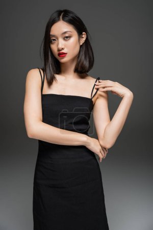 Téléchargez les photos : Charmante femme asiatique en robe noire élégante regardant la caméra sur fond gris - en image libre de droit