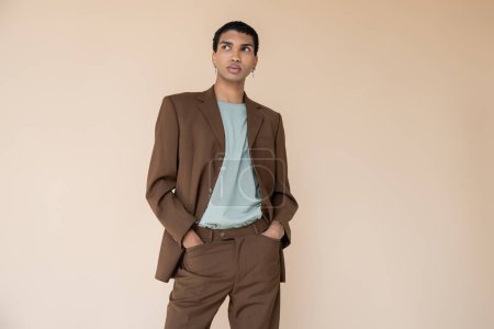 hombre afroamericano de moda en traje marrón cogido de la mano en bolsillos y mirando hacia otro lado aislado en beige