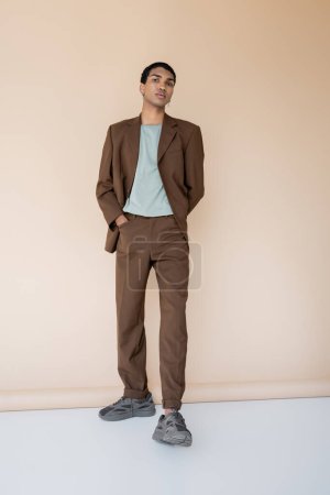 Foto de Longitud completa de hombre afroamericano en traje marrón de pie con la mano en el bolsillo sobre fondo beige - Imagen libre de derechos
