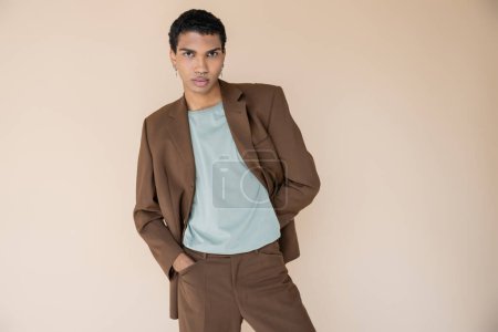 junger afrikanisch-amerikanischer Mann im braunen Anzug steht mit der Hand in der Tasche und blickt isoliert in die Kamera auf beige