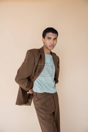 Foto de Hombre afroamericano con estilo en chaqueta marrón mirando a la cámara mientras posando con las manos detrás de la espalda aislado en beige - Imagen libre de derechos