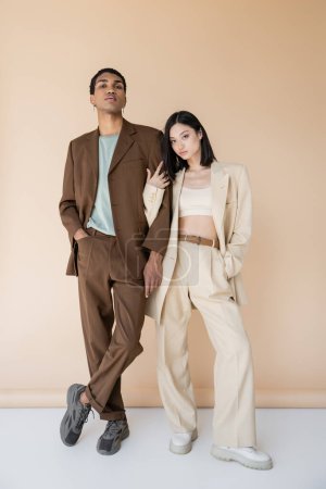 Full length of multiethnic couple in trendy pantsuits posing with hands in pockets on beige background