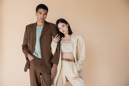 Photo for Young multiethnic couple in trendy pantsuits posing with hands in pockets isolated on beige - Royalty Free Image