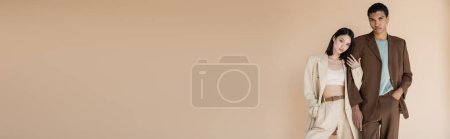 Téléchargez les photos : Jeune couple interracial en costumes tendance debout avec les mains dans les poches isolées sur beige, bannière - en image libre de droit
