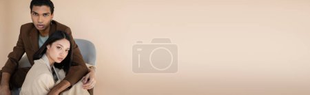 charming asian woman and stylish african american man looking at camera isolated on beige, banner