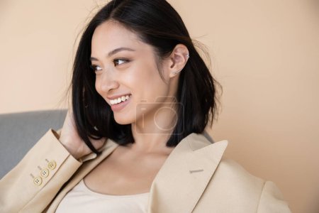 cheerful and stylish asian woman in blazer looking away on beige background