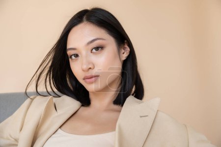 retrato de morena mujer asiática en chaqueta de marfil mirando a la cámara sobre fondo beige