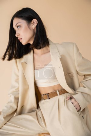 Téléchargez les photos : Brunette asiatique femme en pantalon élégant posant avec la main dans la poche et regardant loin sur fond beige - en image libre de droit