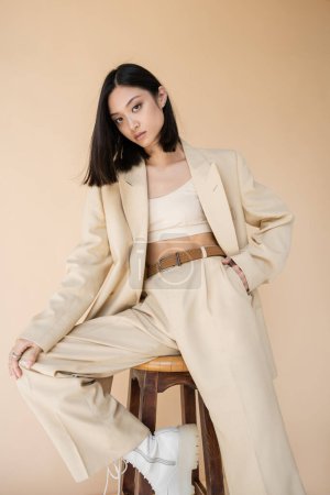 stylish asian woman in pantsuit holding hand in pocket while posing on stool isolated on beige