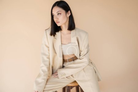 trendy asian woman in ivory suit sitting on stool and looking away isolated on beige