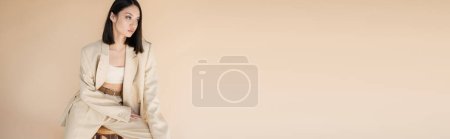 Téléchargez les photos : Brunette asiatique femme en costume ivoire élégant assis sur tabouret et regardant loin isolé sur beige, bannière - en image libre de droit
