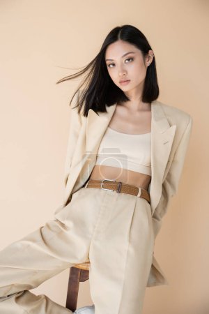 fashionable asian woman in pantsuit posing near stool and looking at camera isolated on beige