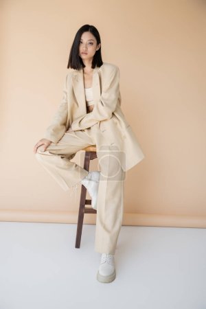 Téléchargez les photos : Brunette asiatique femme en costume élégant et bottes assis sur tabouret sur fond beige - en image libre de droit