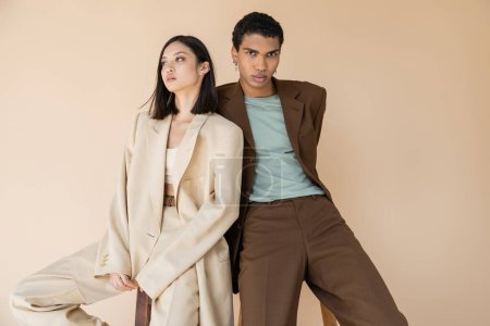 fashionable african american man in brown suit looking at camera near brunette asian model isolated on beige