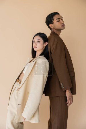 young asian woman in pantsuit standing back to back with trendy african american man and looking at camera isolated on beige