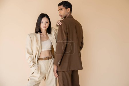 mujer asiática en traje de pantalón de moda mirando a la cámara mientras posa con la mano en el bolsillo cerca de hombre afroamericano aislado en beige
