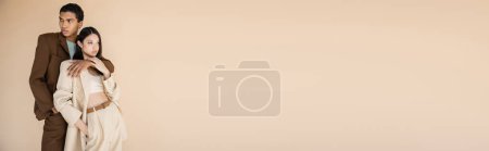 Téléchargez les photos : Jeune homme afro-américain étreignant modèle asiatique en combinaison élégante isolé sur beige, bannière - en image libre de droit