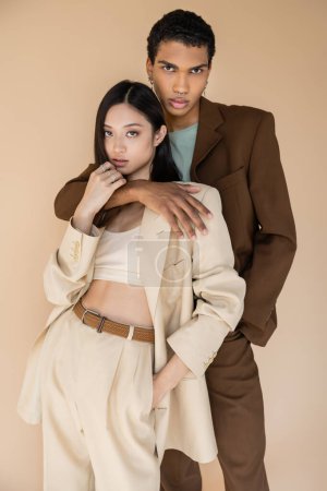 hombre afroamericano de moda abrazando a mujer asiática en traje de pantalón y mirando a la cámara aislada en beige